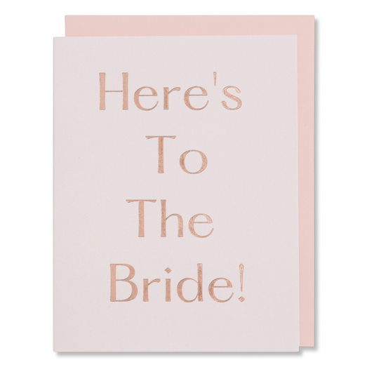 Bridal shower card with rose gold foil on light pink paper, reads 'Here's To the Bride,' elegant and feminine, shown with matching blush envelope