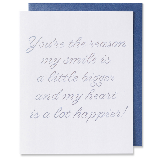 Letterpress romantic love card with You're the reason my smile is bigger and my heart is a lot happier!  On white paper with silver foil stamped. Metallic blue envelope